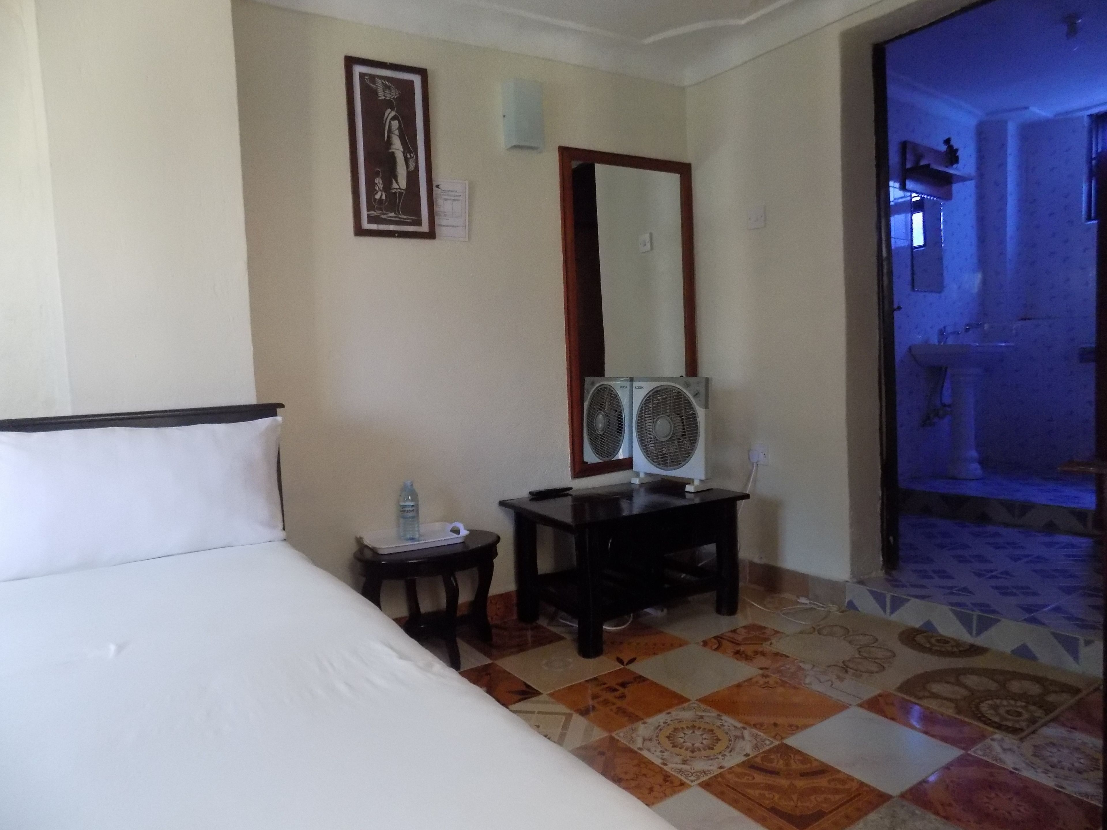 A hotel room with a bed, a small table with a water bottle, a mirror, and two wall fans.