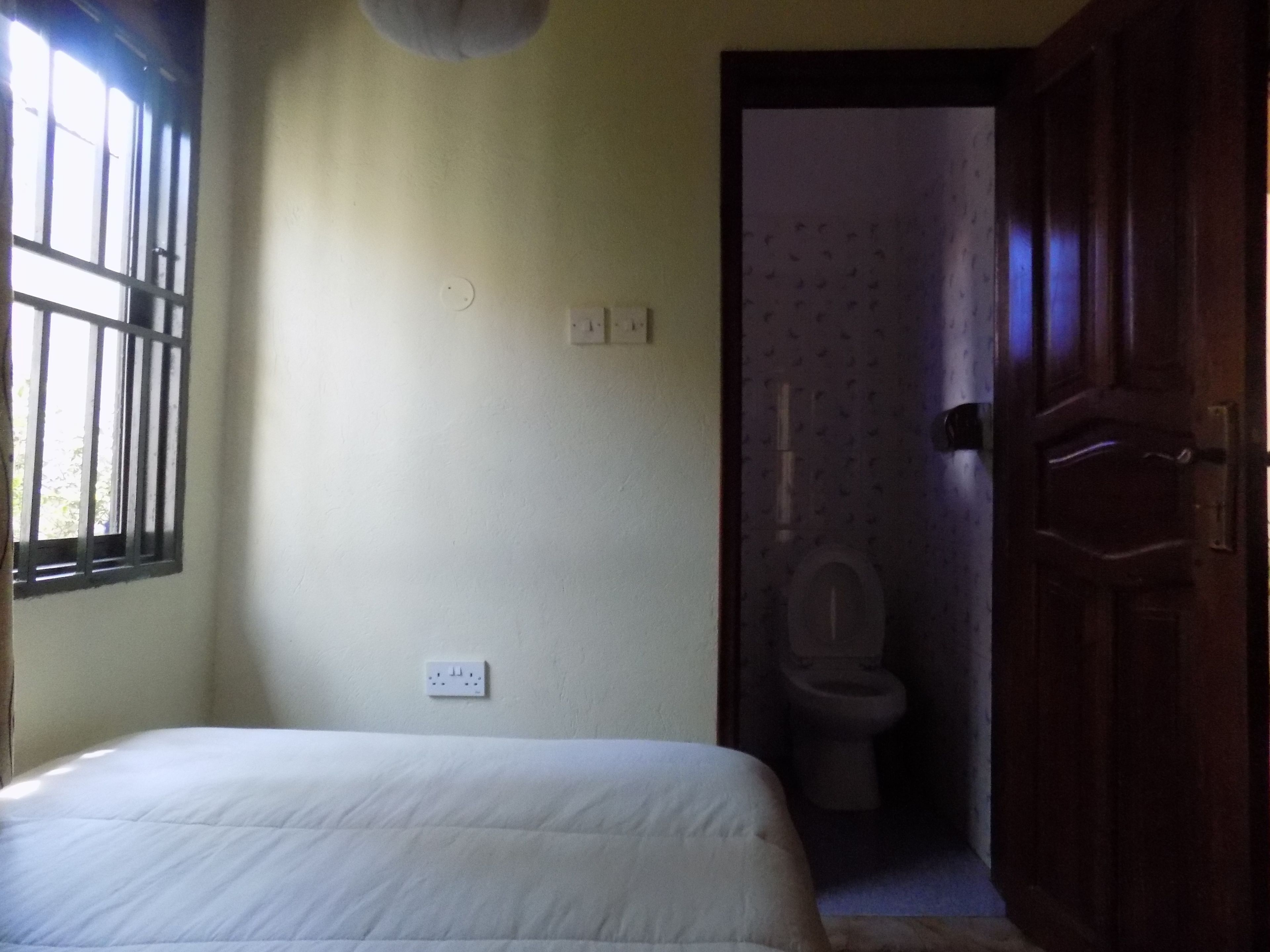 A bedroom with a bed, a window with bars, a door leading to a bathroom, and a light switch on the wall.
