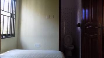 A bedroom with a bed, a window with bars, a door leading to a bathroom, and a light switch on the wall.