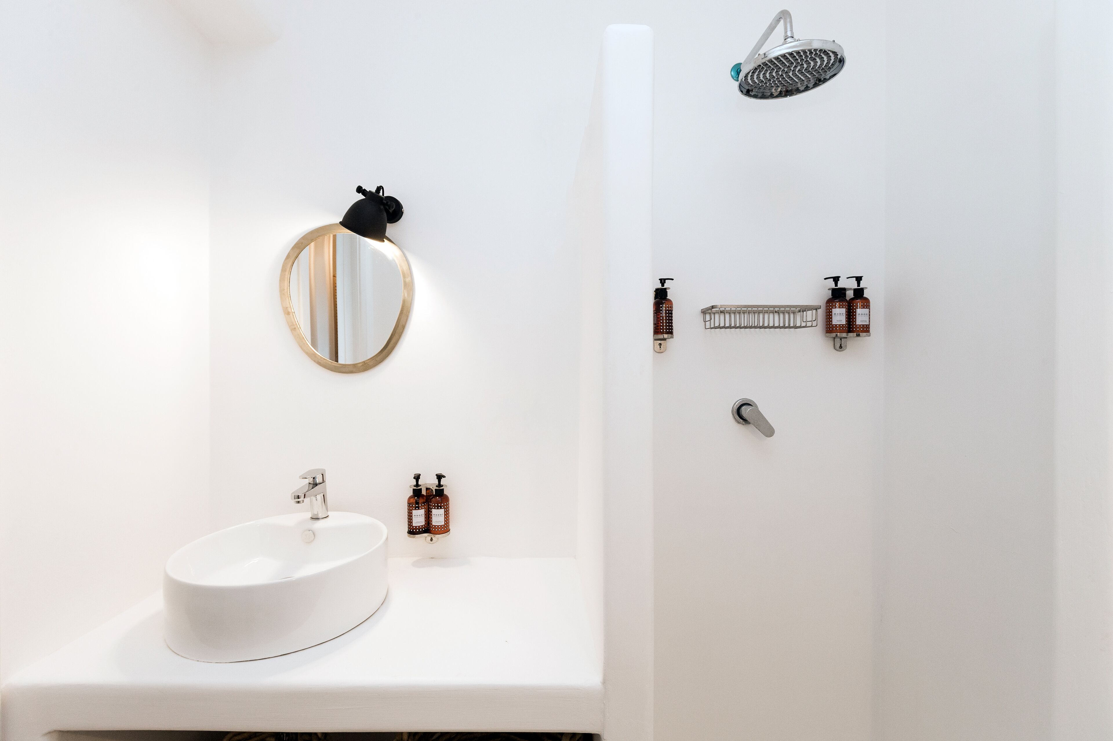 A modern bathroom with a white sink, a round mirror with a gold frame, a handheld showerhead, and two bottles of soap.