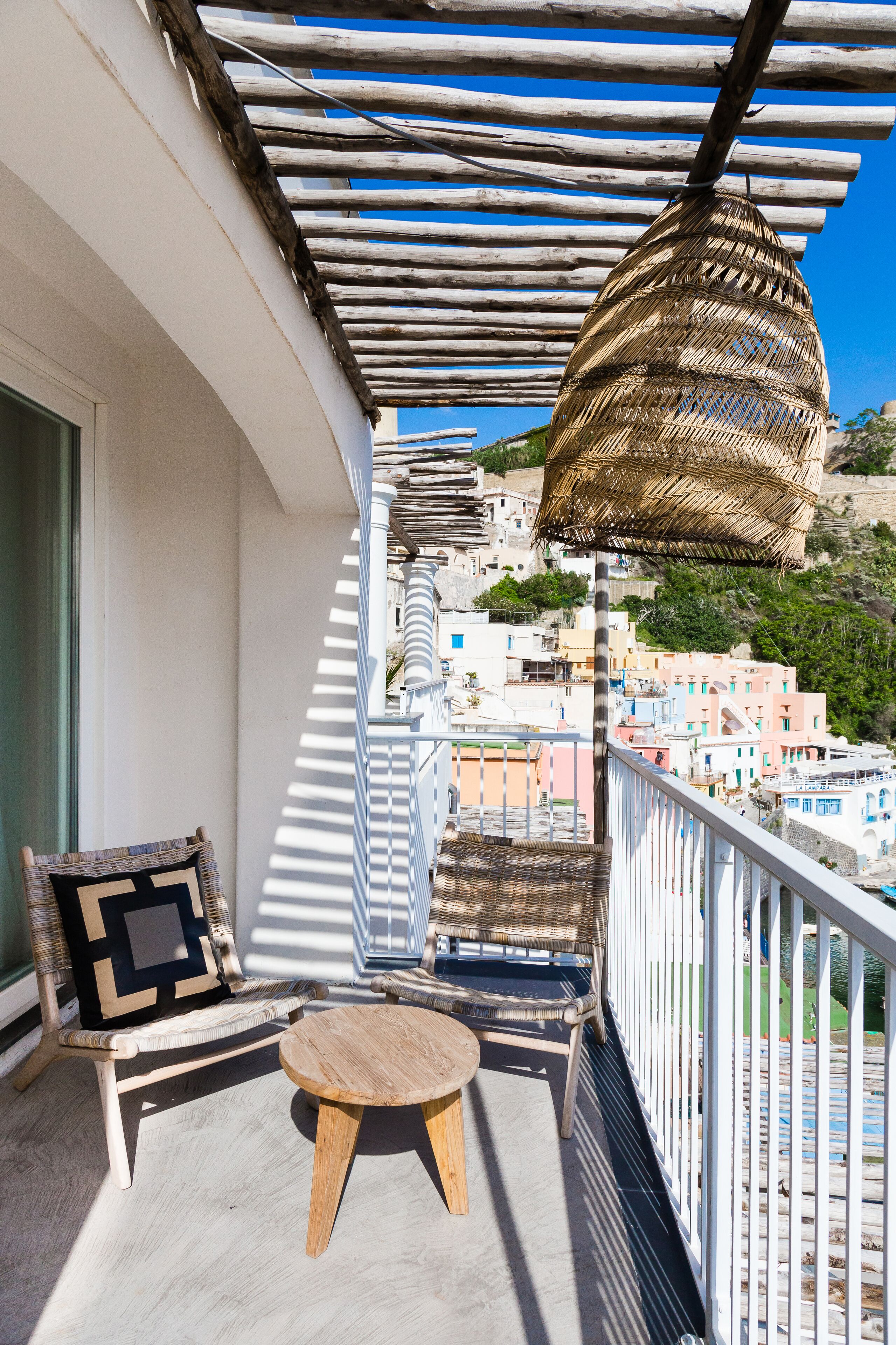 A balcony with wicker furniture, a hanging basket, and a view of a coastal town.