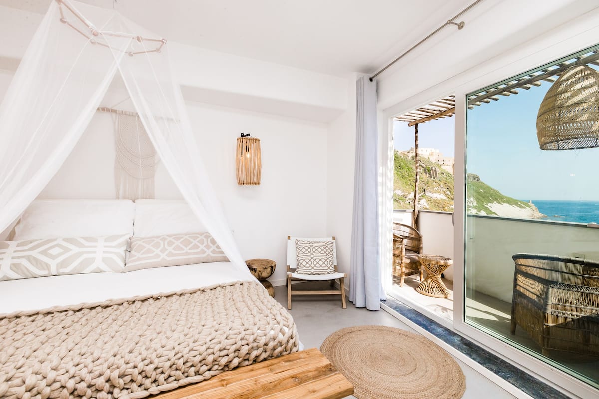A bedroom with a bed, a chair, a woven rug, and a view of the sea through a glass door.