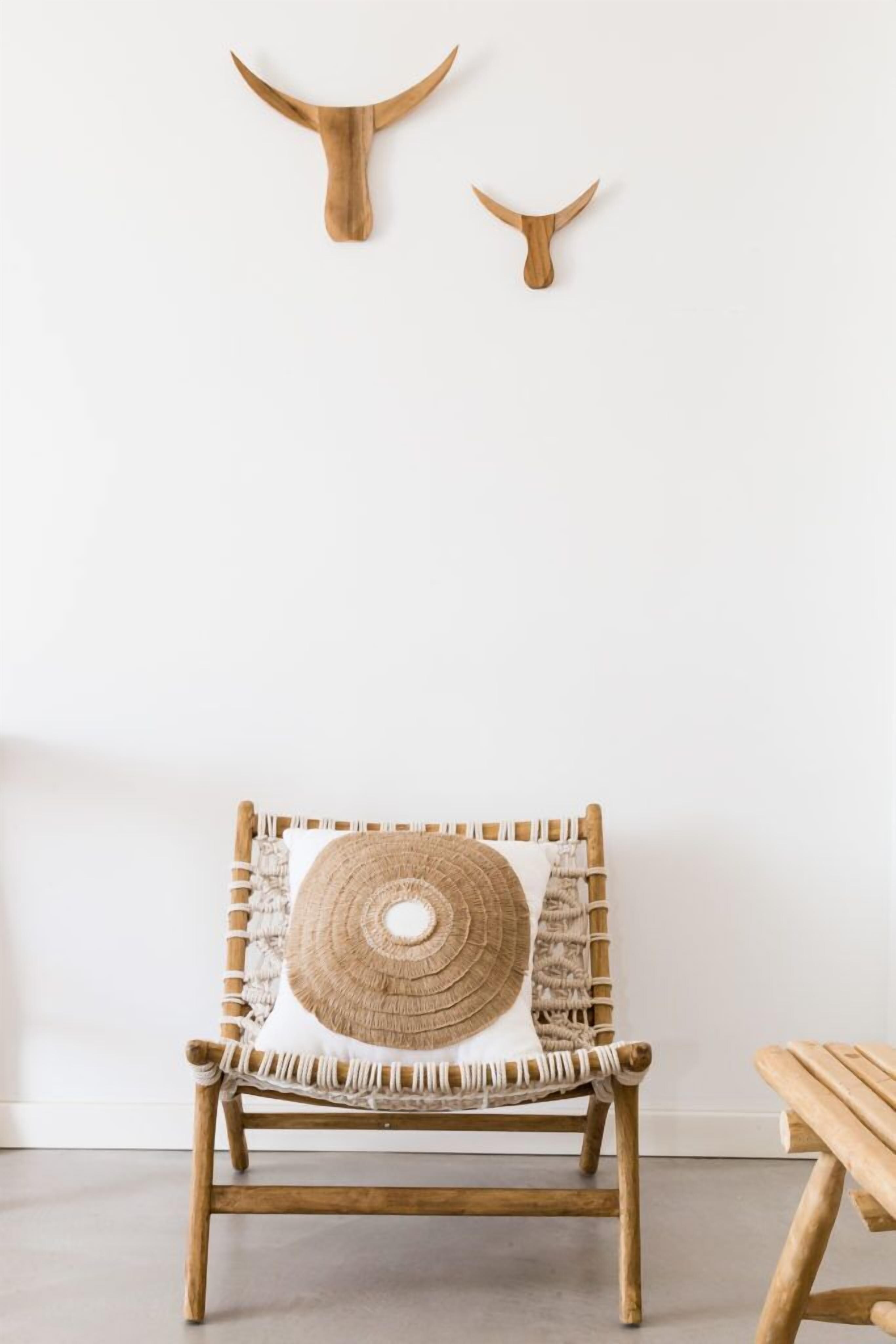Una silla de madera con respaldo tejido y cojín circular tejido, ubicada frente a una pared blanca con dos adornos de madera en forma de cuerno.