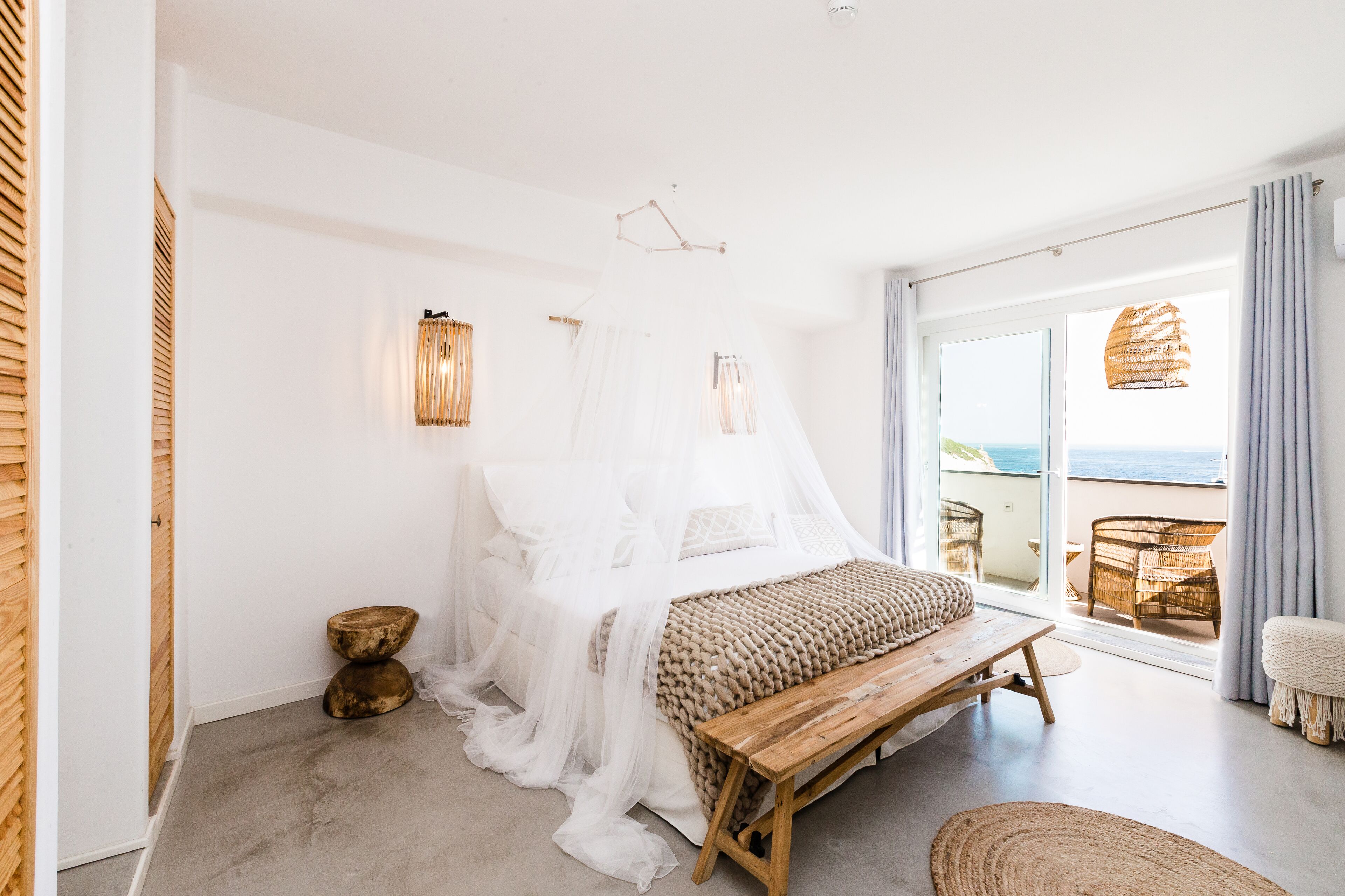 Un dormitorio con una cama cubierta de sábanas blancas, un banco de madera, una silla de mimbre y vistas al mar a través de amplias puertas correderas.