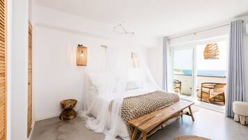 Un dormitorio con una cama cubierta de sábanas blancas, un banco de madera, una silla de mimbre y vistas al mar a través de amplias puertas correderas.