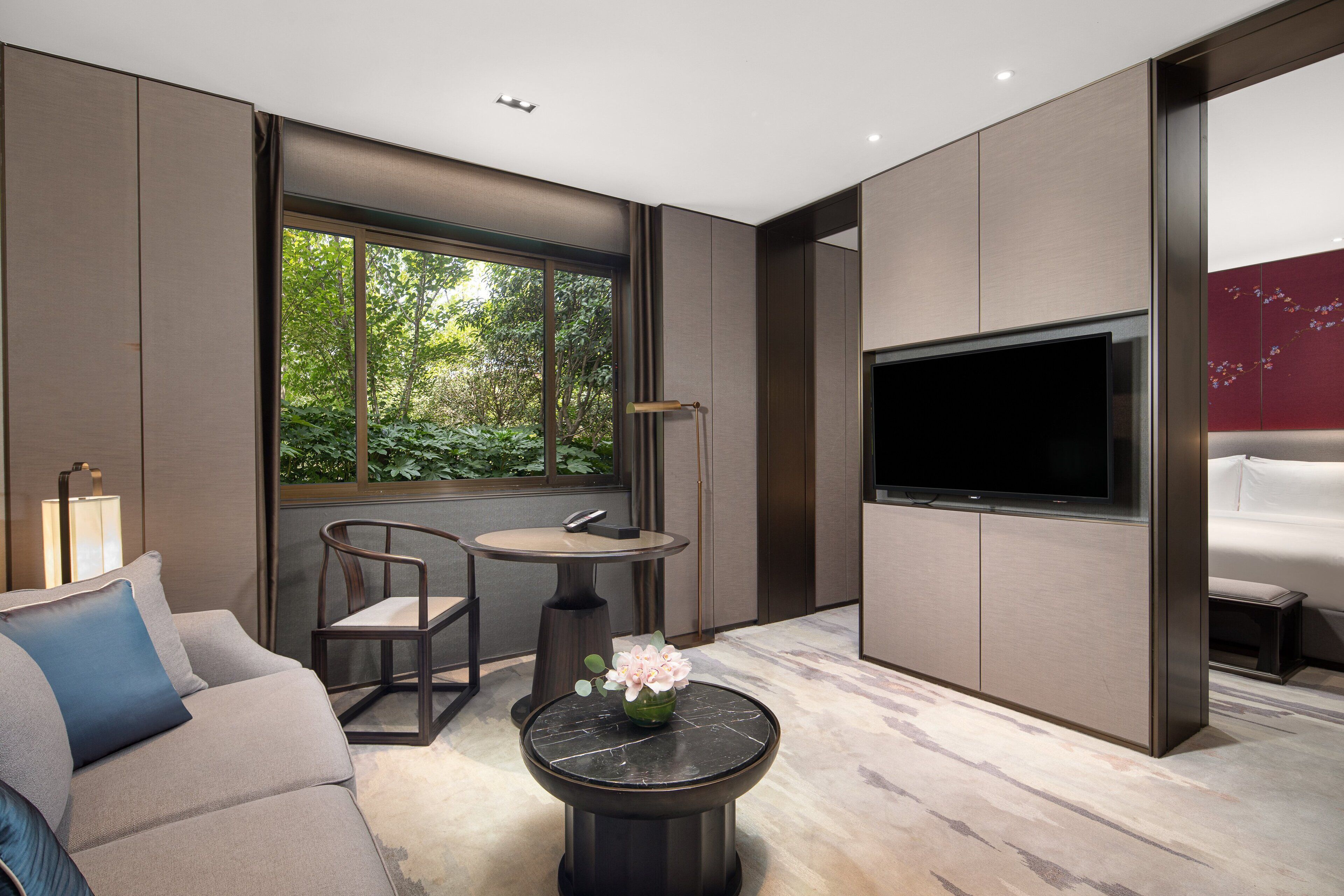 Una sala de estar moderna con un sofá, una mesa de centro redonda con un florero, una mesa de comedor con sillas, un televisor montado en la pared y un amplio ventanal con vistas a zonas verdes.
