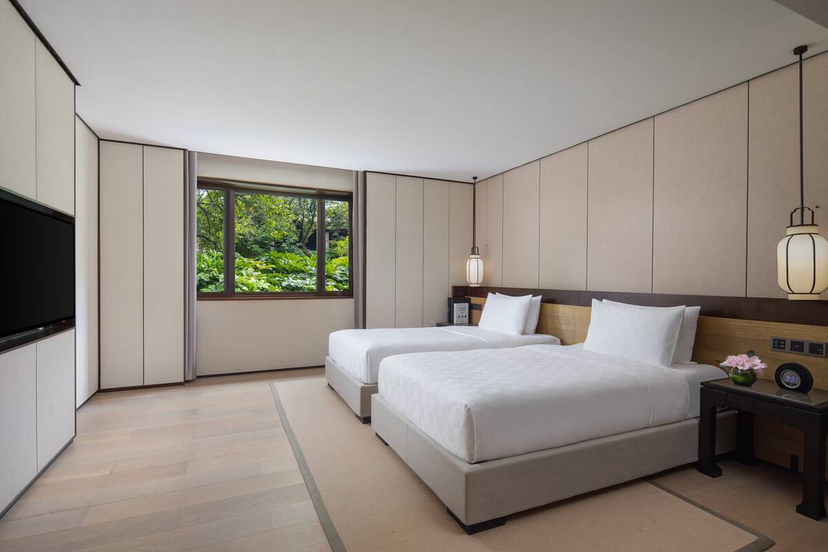 Habitación de hotel moderna con dos camas, televisor de pantalla plana, mesita de noche con lámpara y un ventanal con vistas a zonas verdes.