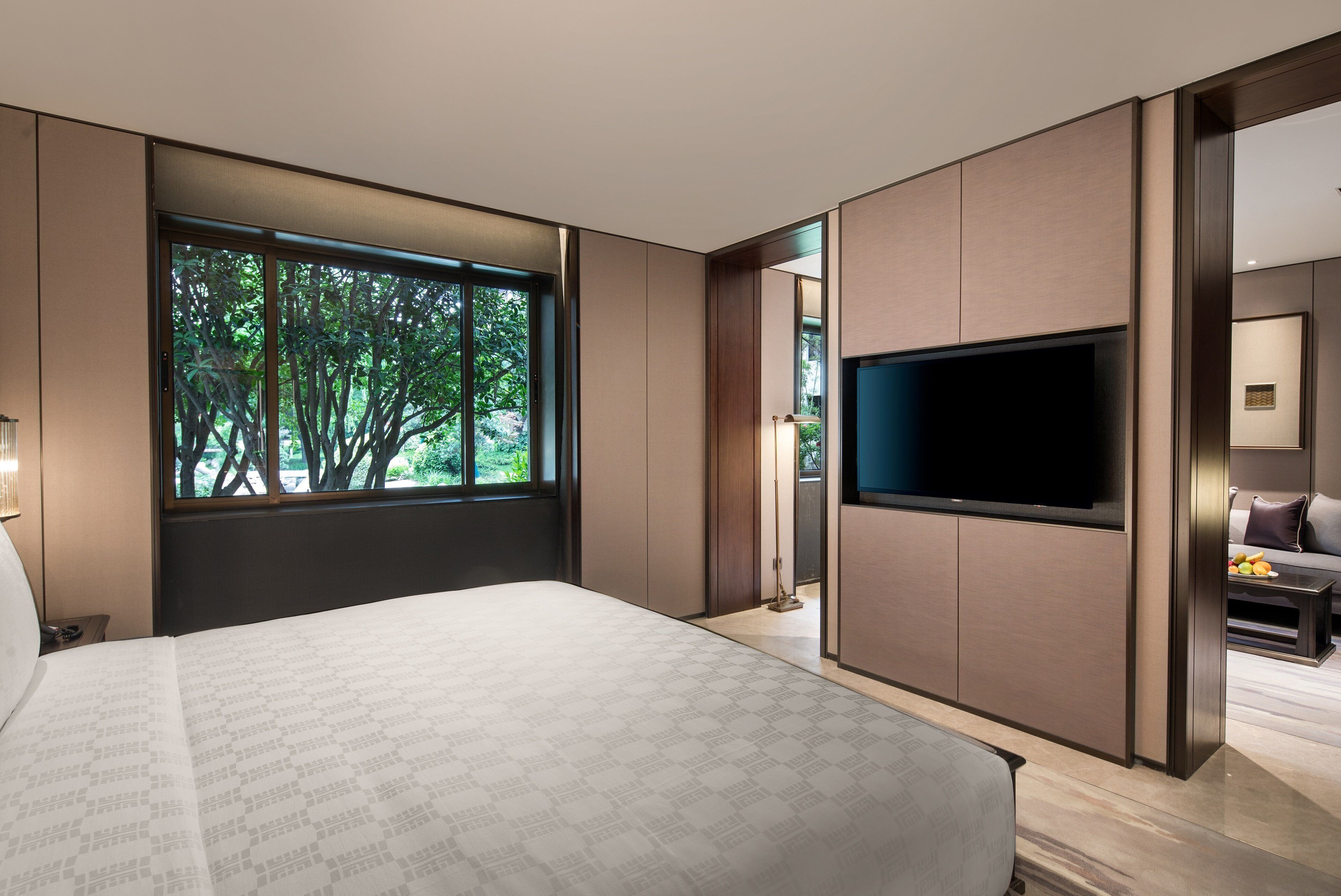 A modern bedroom with a large window, a flat-screen TV, and a view of trees.