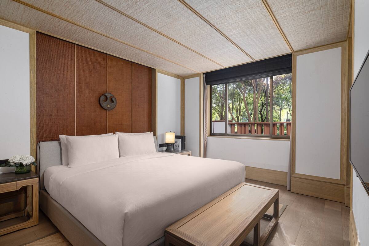 Habitación de hotel con una cama grande, una mesita de noche de madera, una mesita con lámpara y una ventana con vistas a árboles.