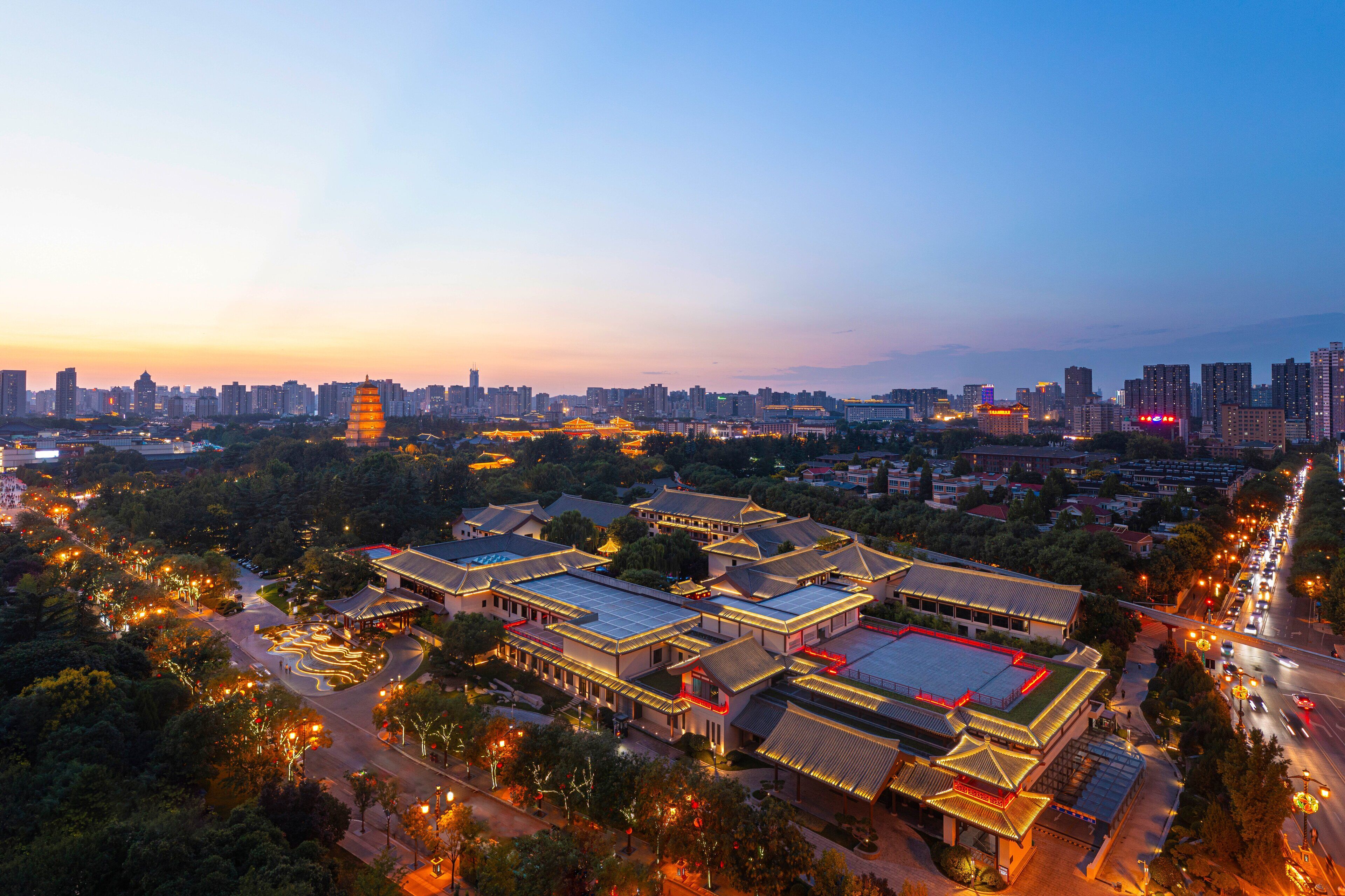 A traditional Chinese architectural complex with multiple buildings, surrounded by greenery and modern cityscape in the background.