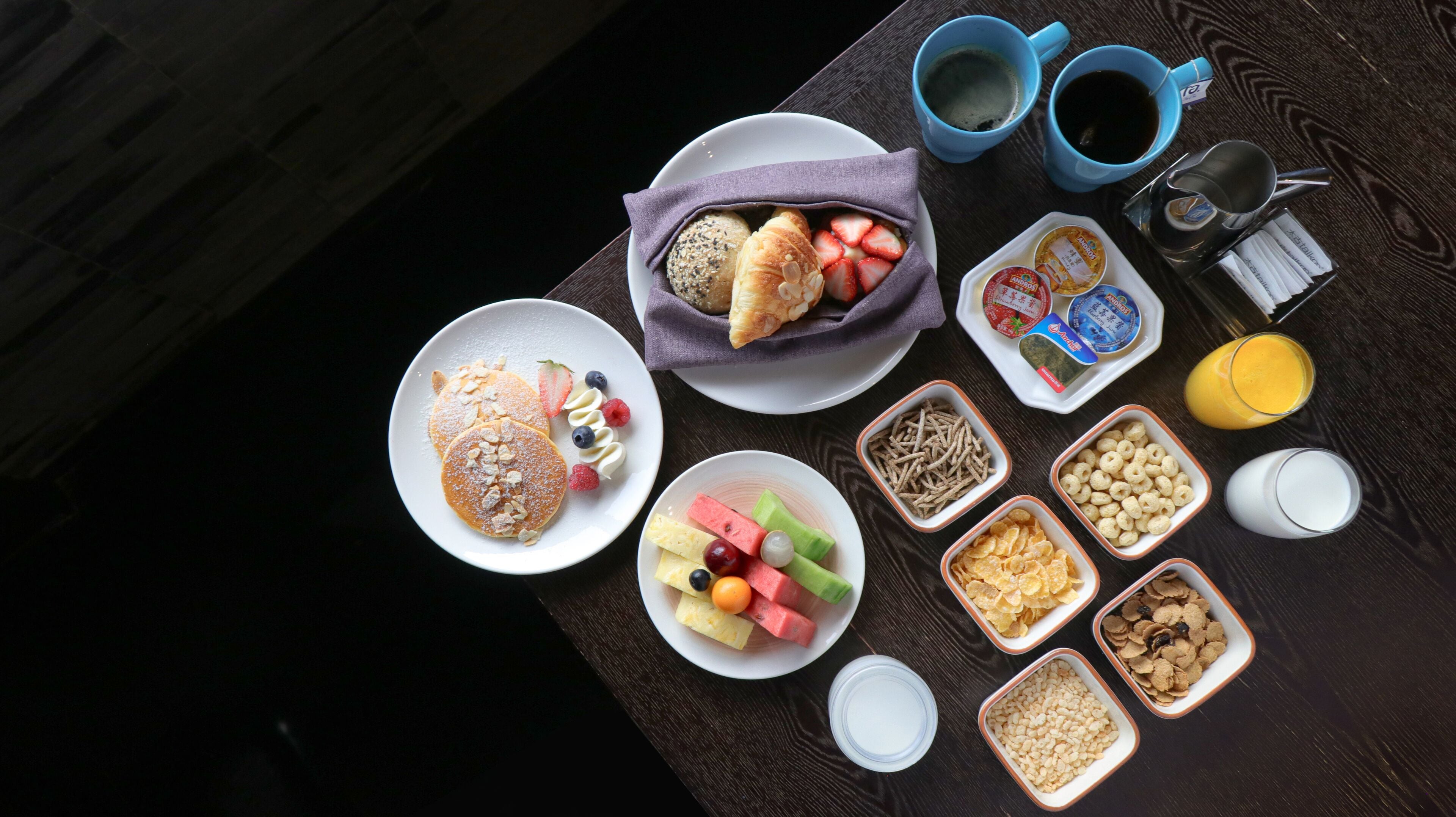 A breakfast table with pancakes, croissants, yogurt, fruit, cereal, and coffee.