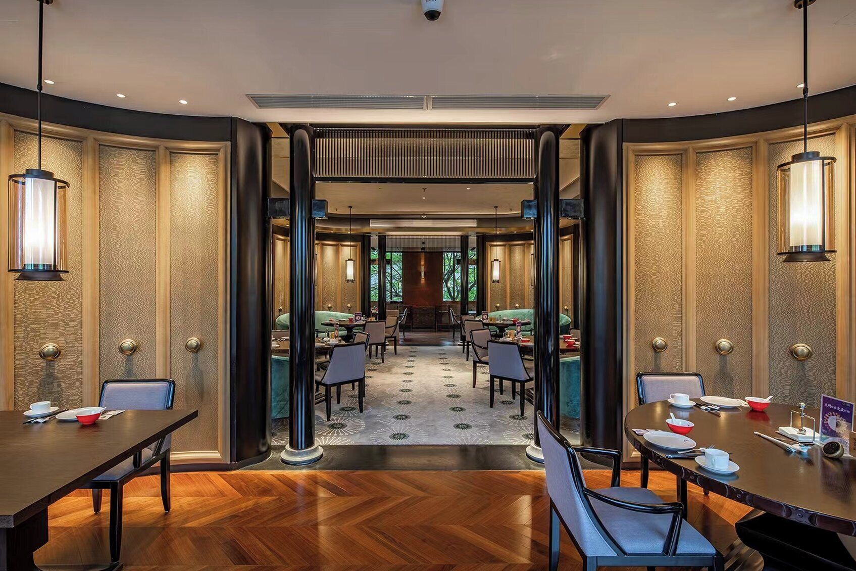 A modern hotel dining area with wooden flooring, round tables, and chairs. There are two large windows providing natural light.