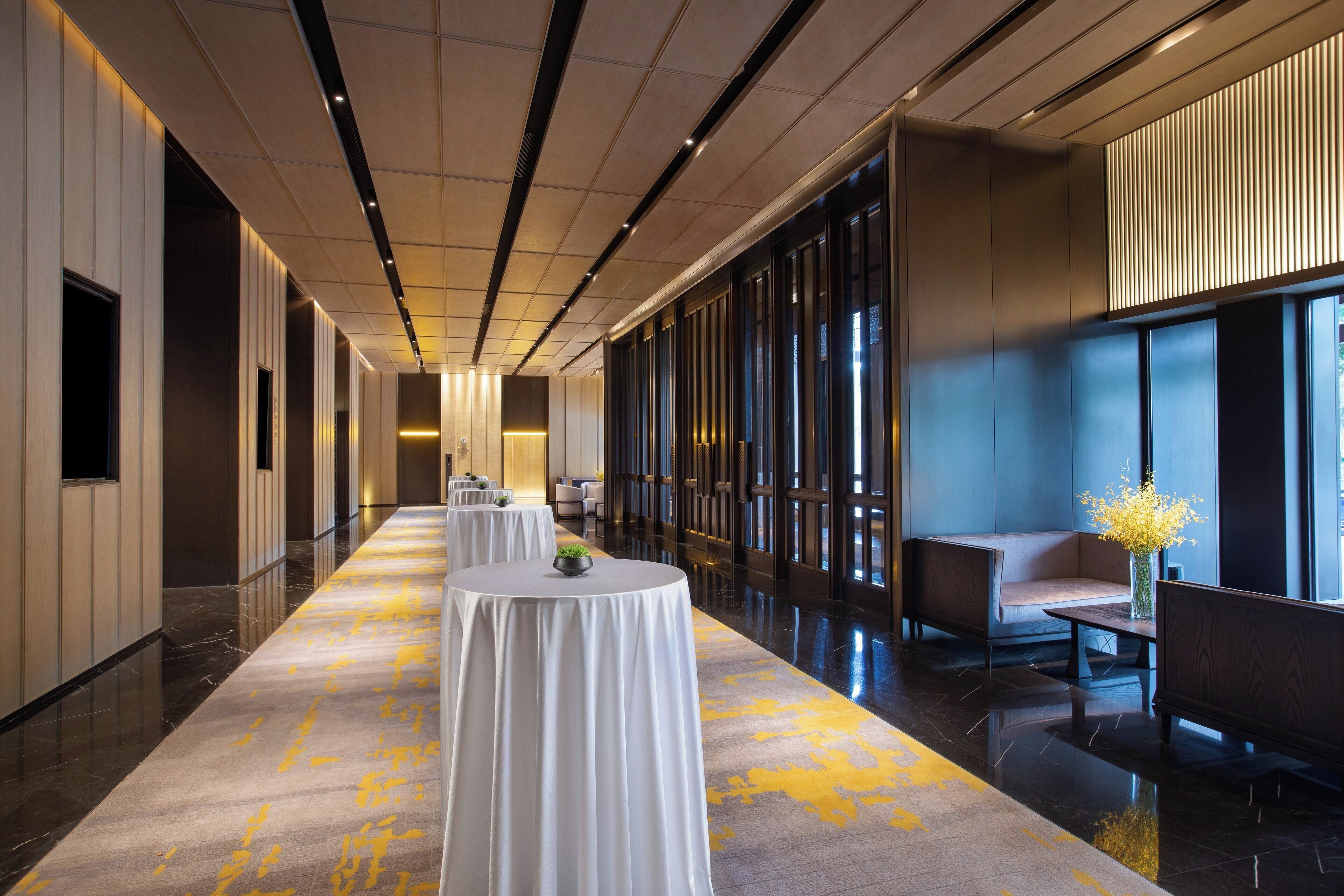 A hotel corridor with round tables, seating areas, and large windows.