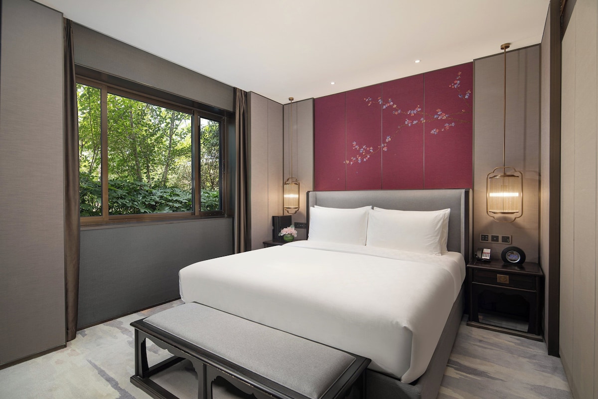 A modern hotel room with a large bed, bedside lamps, a small table, and a view of greenery through the window.