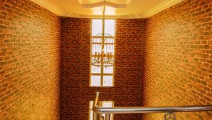 Un escalier étroit aux murs de briques, une rampe métallique et un lustre suspendu au plafond.