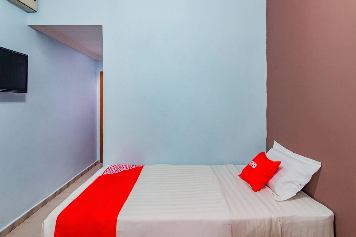 Een slaapkamer met een bed, een rood-wit dekbed, een rood kussen en een televisie aan de muur.