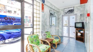 Un hall d’hôtel avec un comptoir de réception, deux fauteuils en osier avec des coussins verts, un téléviseur posé sur une étagère en bois et une grande fenêtre donnant sur les voitures garées.