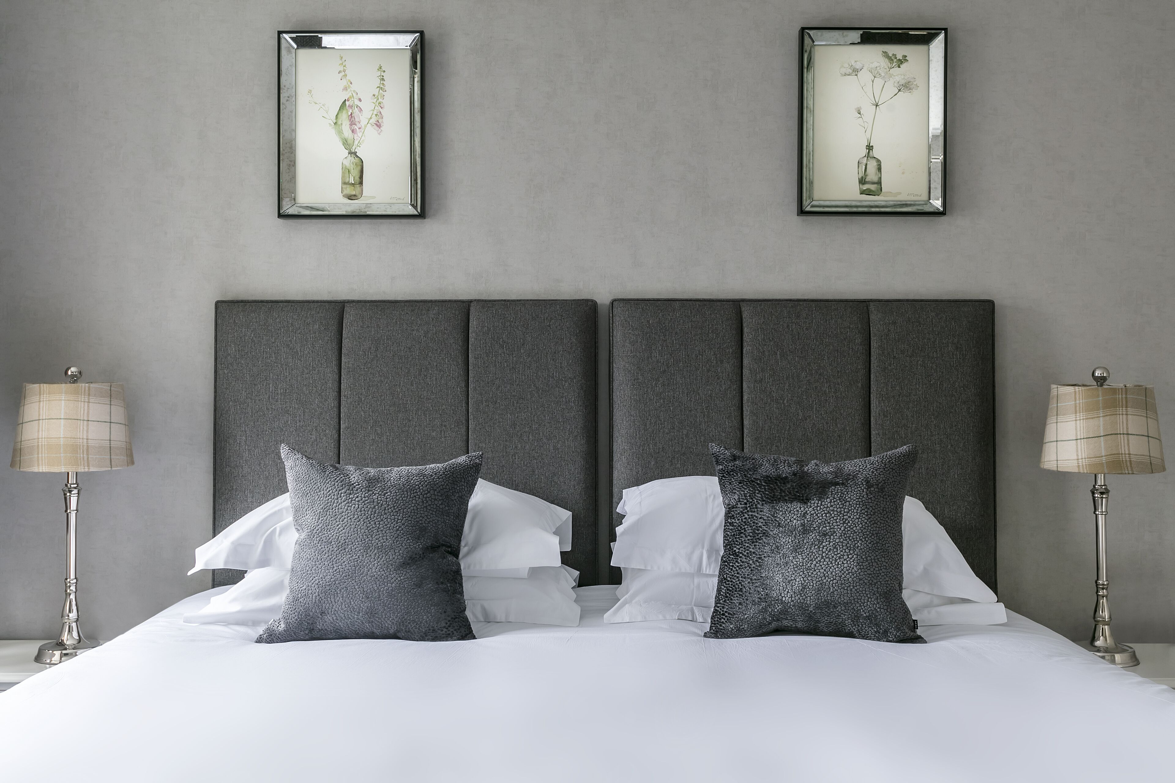 Uma cama de casal com cabeceira cinza e roupa de cama branca, com duas mesas de cabeceira com luminárias, e duas obras de arte com flores emolduradas na parede.