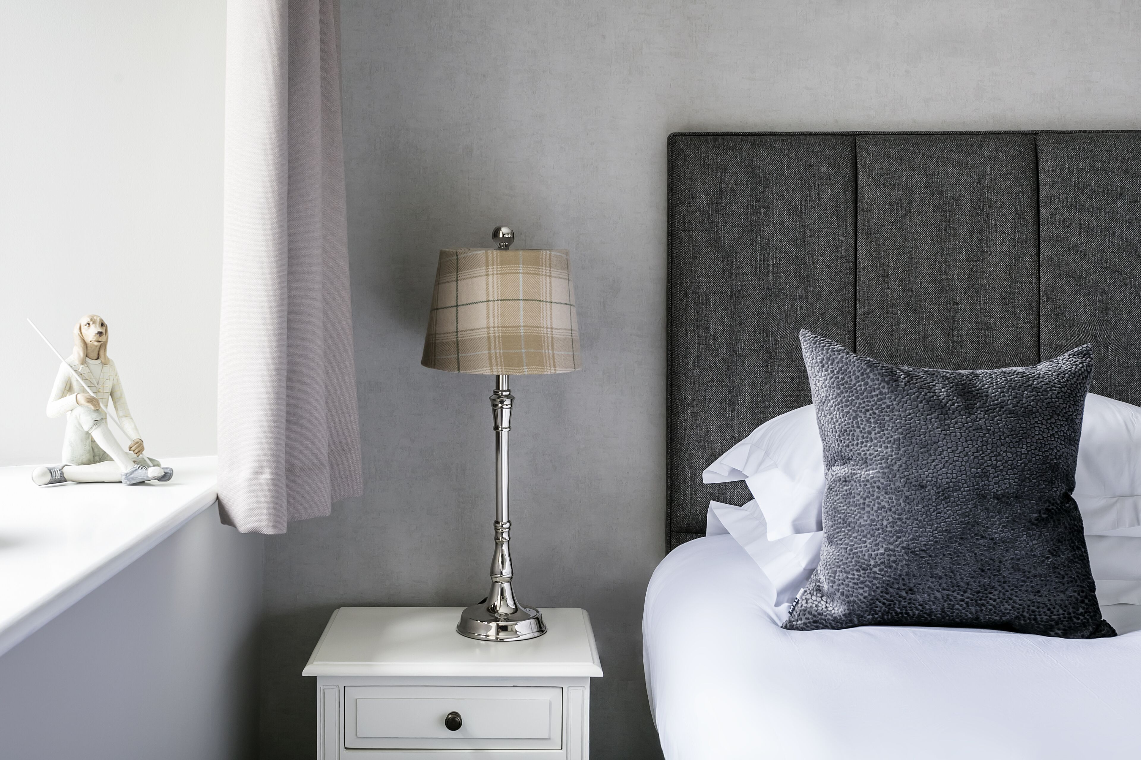 A neatly made bed with a grey pillow, a white nightstand with a lamp, and a small figurine on the nightstand.