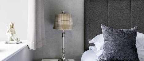 A neatly made bed with a grey pillow, a white nightstand with a lamp, and a small figurine on the nightstand.