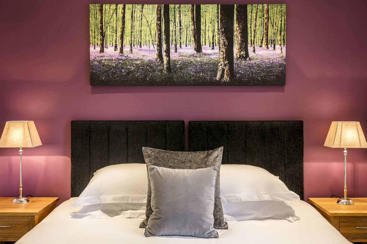 A bedroom with a large bed, two bedside tables with lamps, a headboard, and a forest-themed painting on the wall.