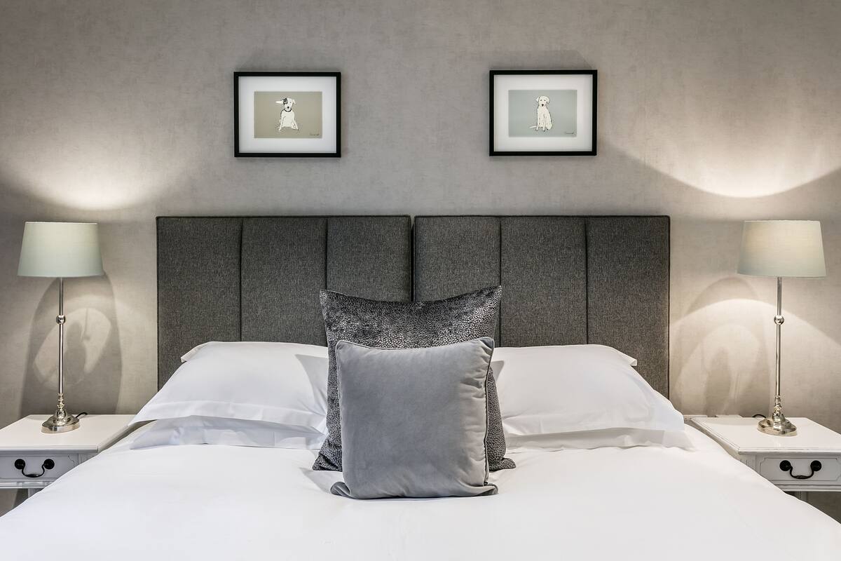 A neatly made bed with white linens and two pillows, flanked by nightstands with lamps, in a room with a grey textured wall and framed artwork above.