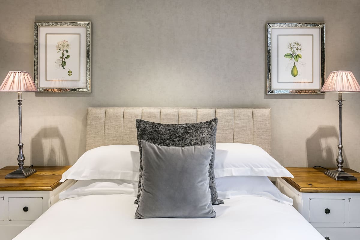 A neatly made bed with a grey pillow, white linens, and a wooden headboard. There are two framed botanical prints on the wall above the bed.