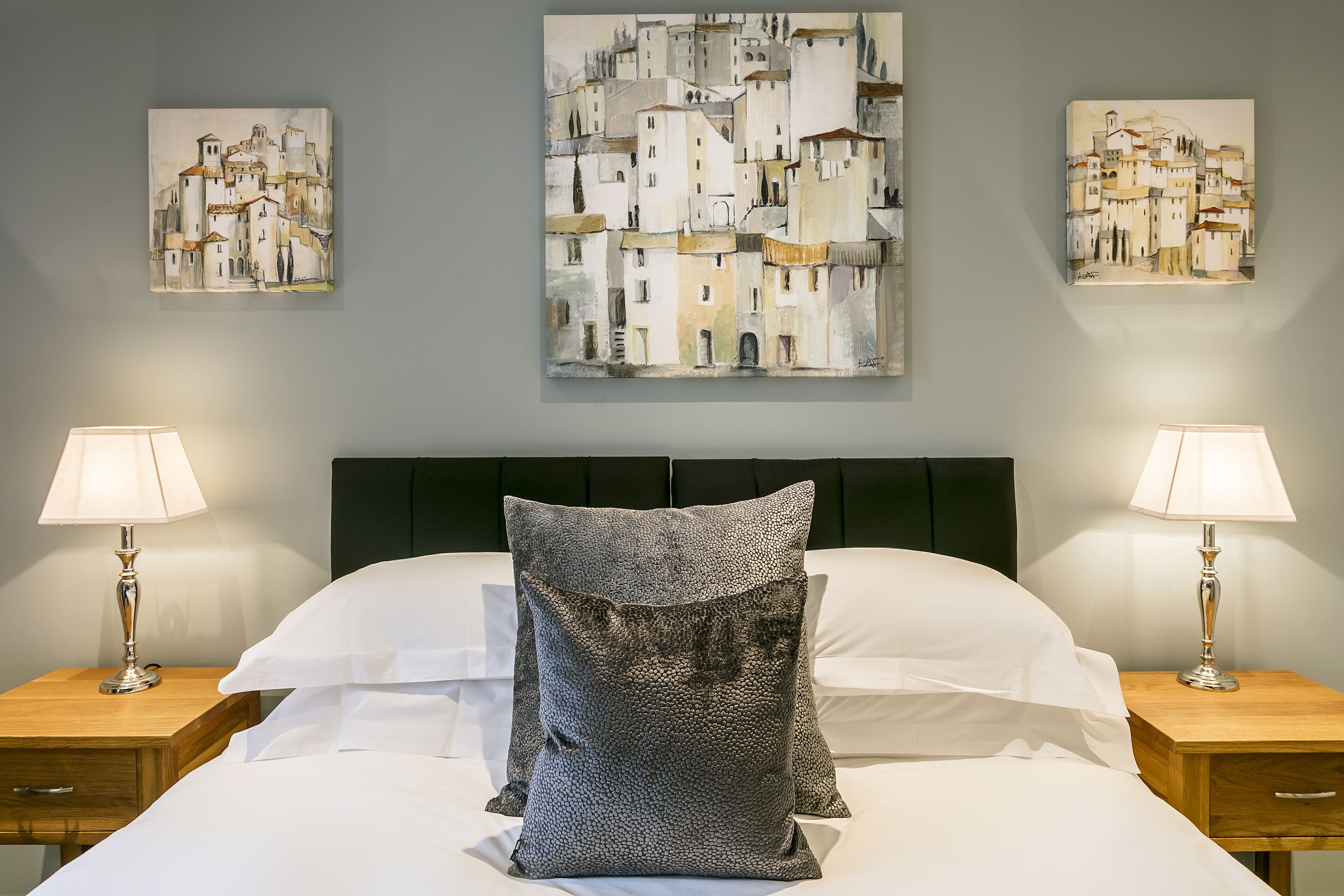A hotel room with a bed, two bedside tables, two lamps, and three paintings on the wall.