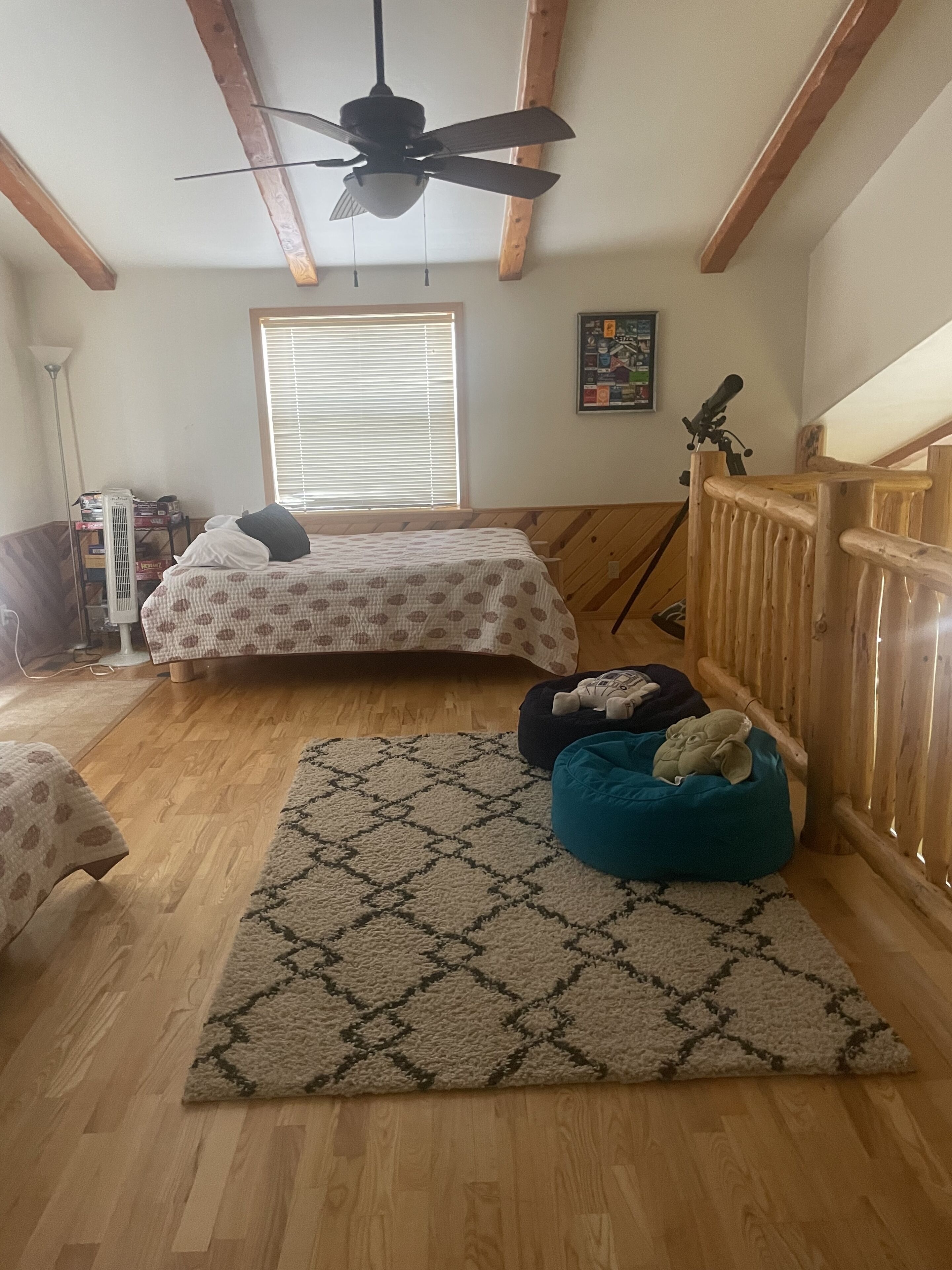 A bedroom with two beds, a ceiling fan, wooden beams, a window with blinds, a floor lamp, and a rug.