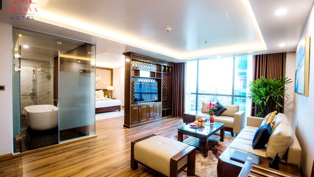 A modern hotel room with a glass-enclosed bathroom, a flat-screen TV, a sofa, and a coffee table. - Menrva Hotel (Hai Phong)