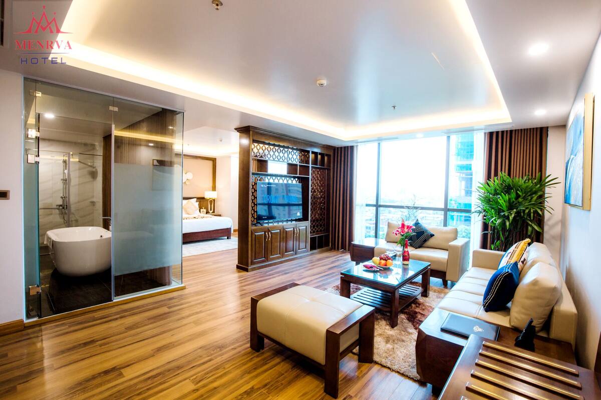 A modern hotel room with a glass-enclosed bathroom, a flat-screen TV, a sofa, and a coffee table.