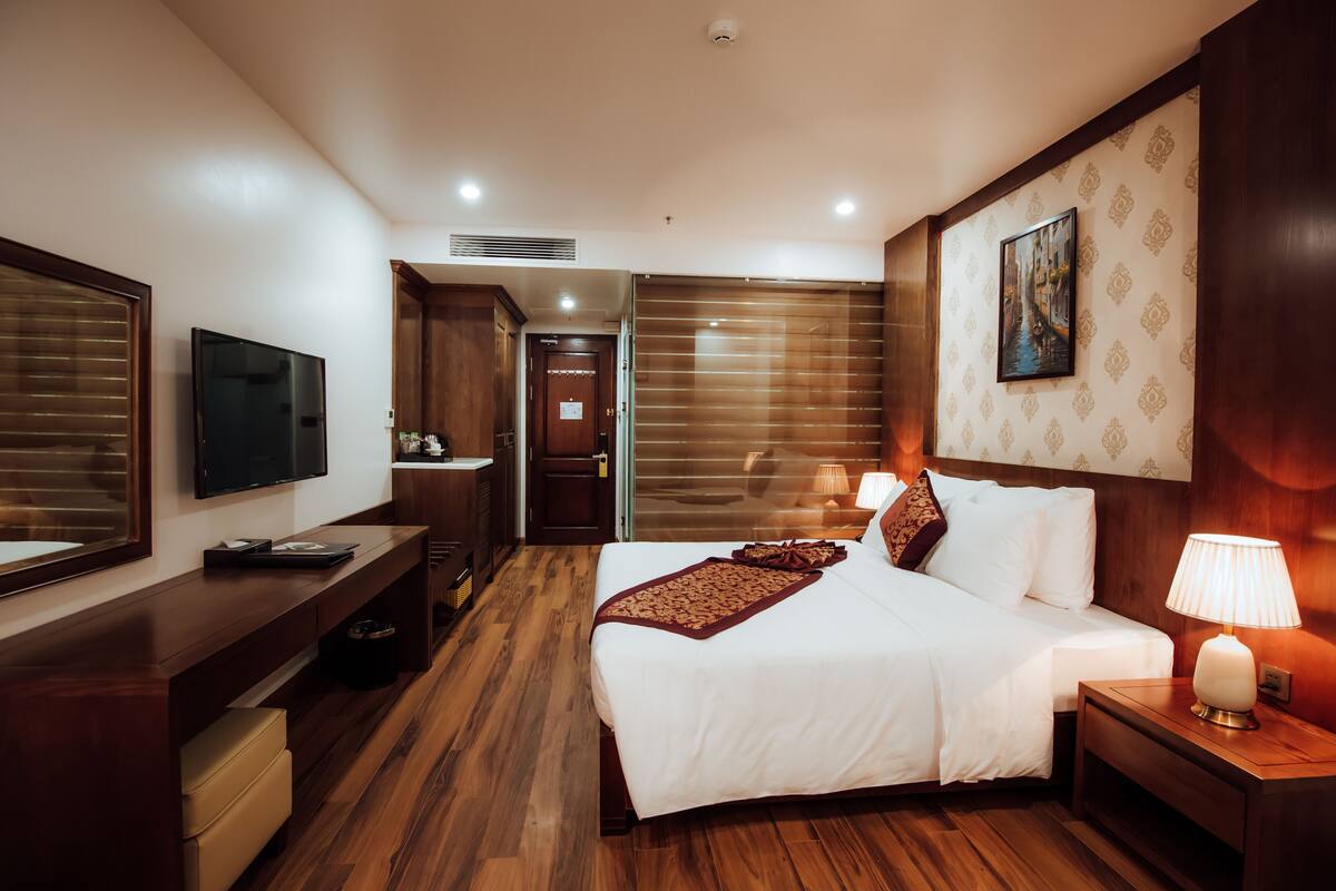 A hotel room with a large bed, a TV, a desk, and a nightstand with lamps.