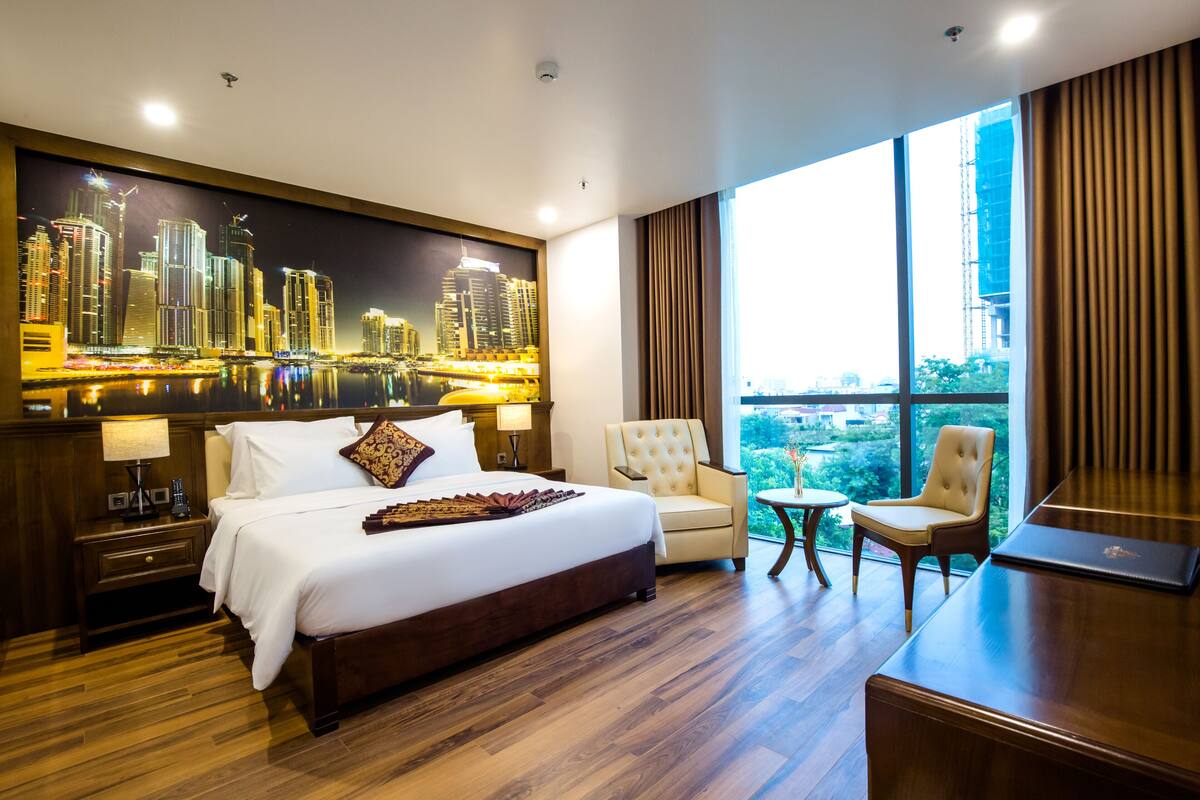 A hotel room with a large bed, two armchairs, a desk, and a cityscape view.