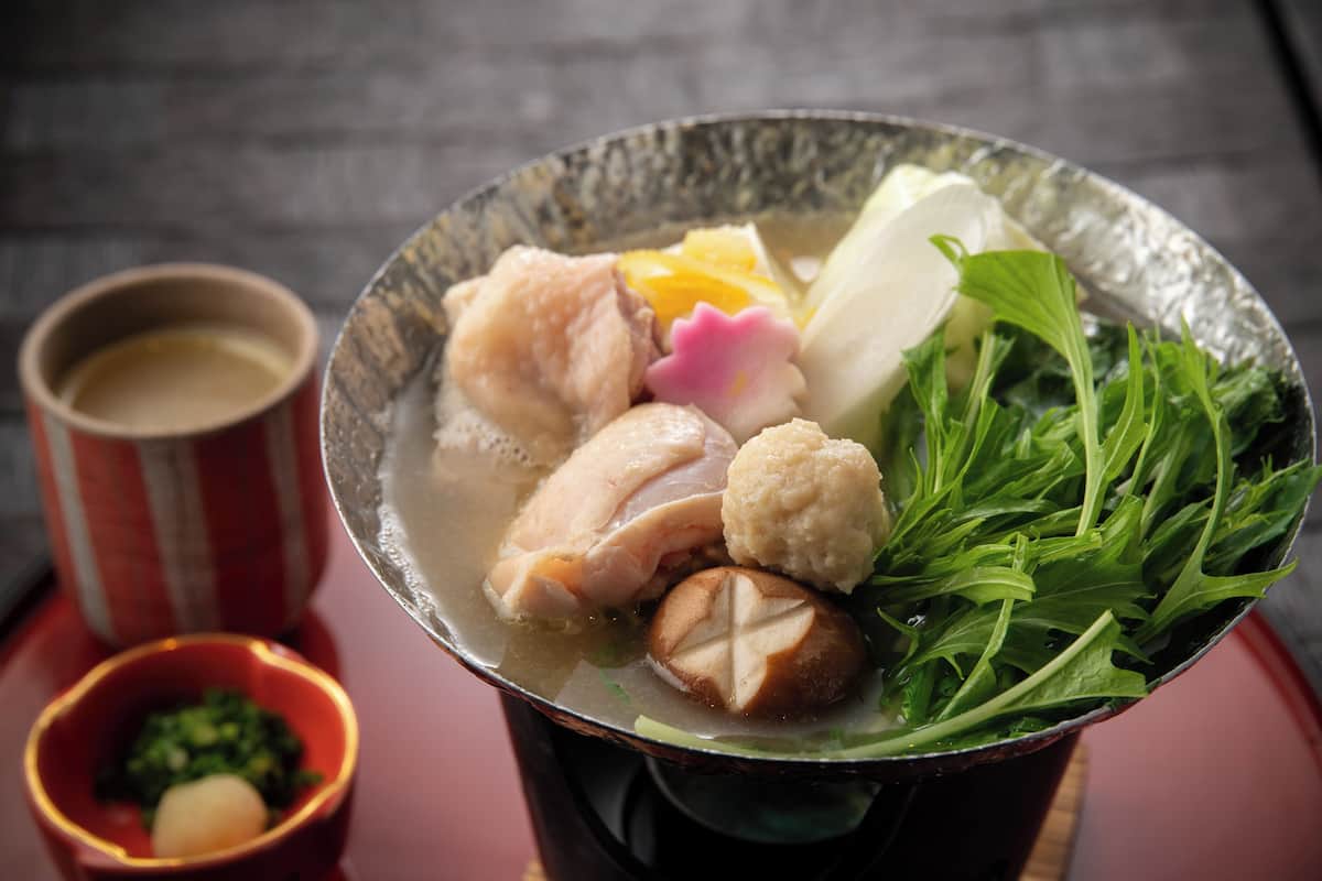 A bowl of soup with various ingredients, a cup of tea, and a small dish of greens.