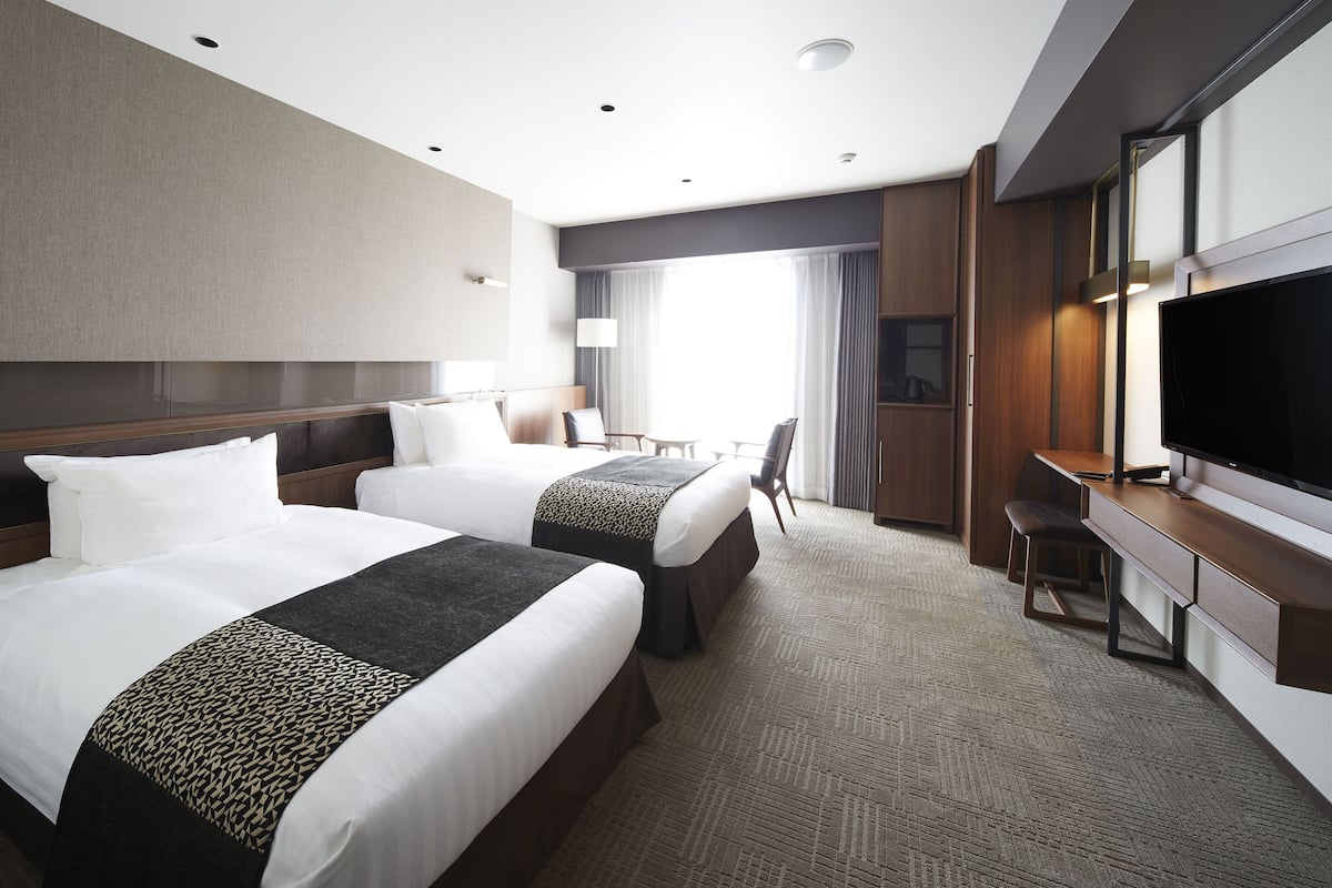 A hotel room with two beds, a desk, a TV, and a large window with curtains.