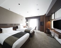 A hotel room with two beds, a desk, a TV, and a large window with curtains.