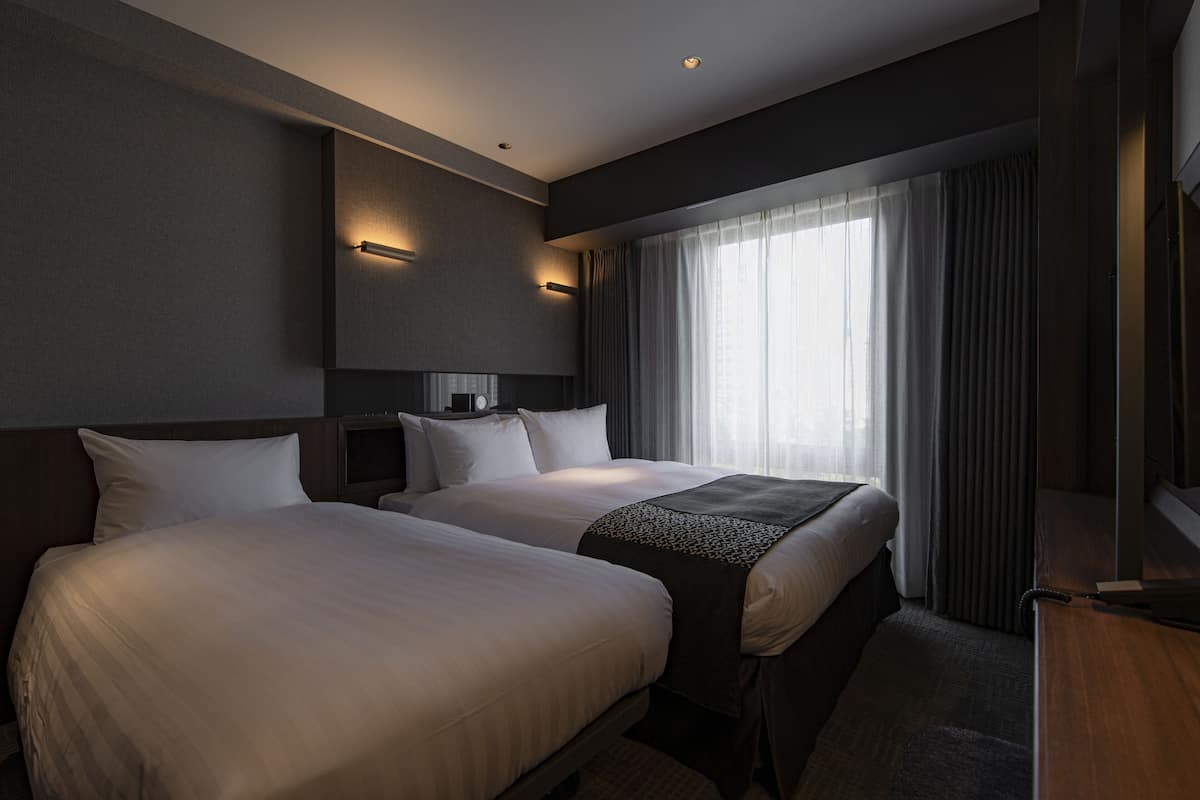 A hotel room with two beds, a large window with curtains, and wall-mounted lighting.