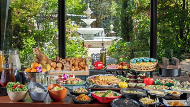 A buffet table with various food items and a view of a fountain through large windows. - Hotel Trad Hakata (Fukuoka)