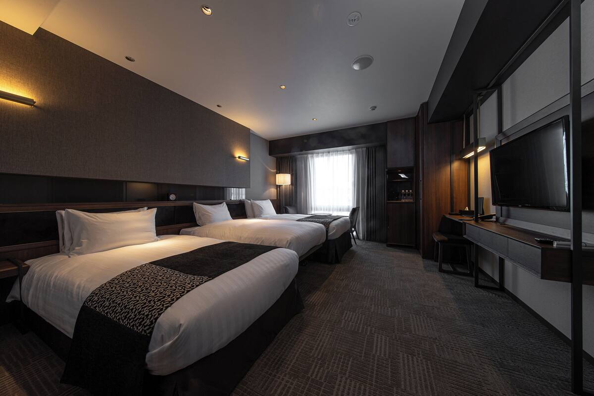A hotel room with two beds, a desk with a chair, a TV, and a large window with curtains.