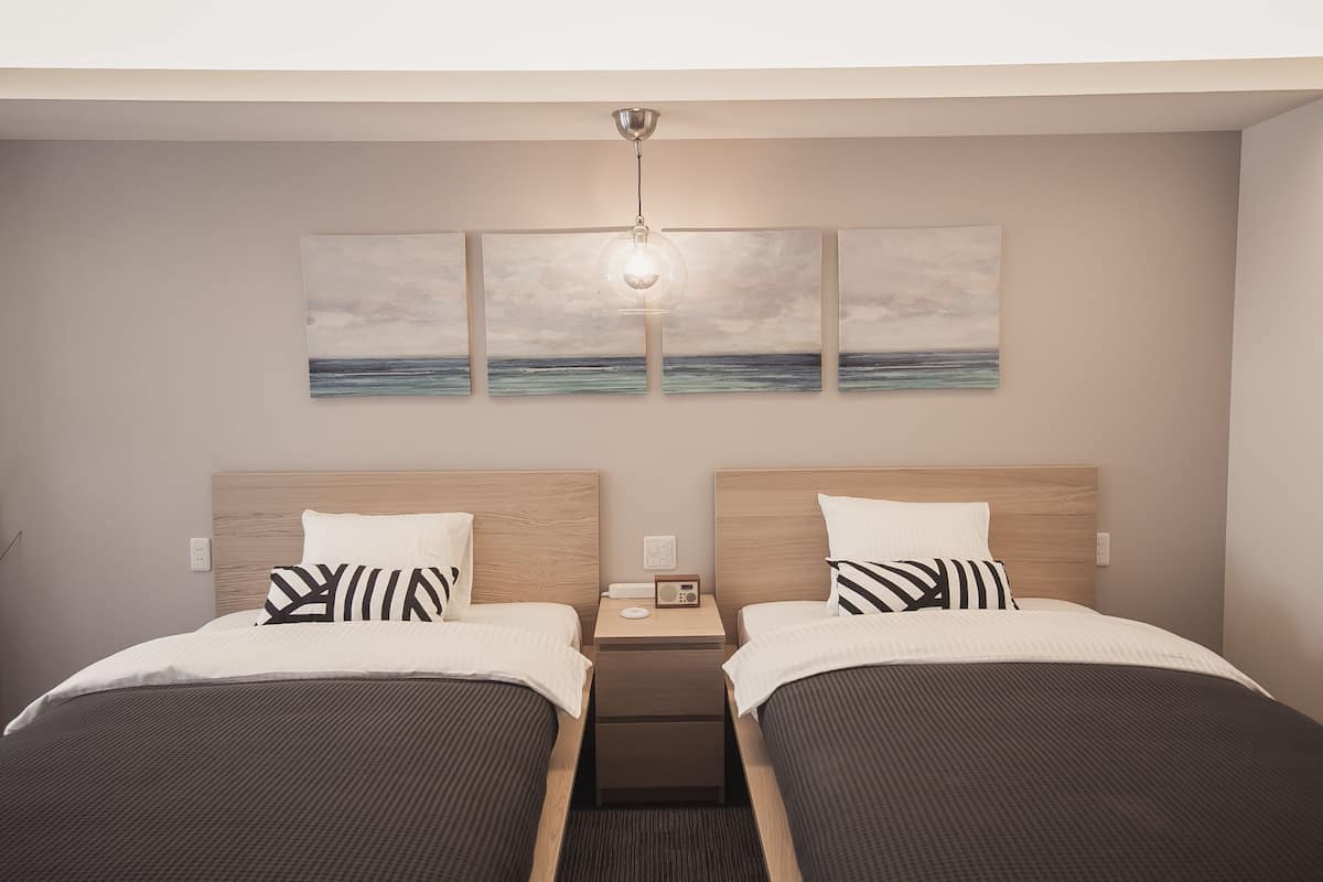 Quarto de hotel com duas camas, uma mesa de cabeceira com rádio e uma obra de arte na parede retratando uma cena de praia.