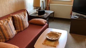 A cozy room with a red sofa, a small table with a woven basket, a lamp, a TV, and a window with curtains.