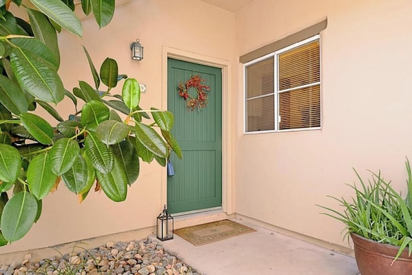A house entrance with a green door, a wreath, a window with blinds, and a potted plant.