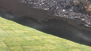 Ein grasbewachsenes Gelände mit einer Steinmauer und felsigem Hintergrund.
