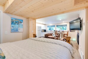 A bedroom with a large bed, a painting of a beach scene, a kitchenette, and a TV mounted on the wall. - 2 Mi to St Edward State Park: Ground-Level Studio (Kenmore)