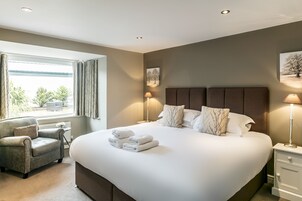 A neatly made bed with white linens and a set of folded towels, a grey armchair, a bedside table with a lamp, and a window with curtains.