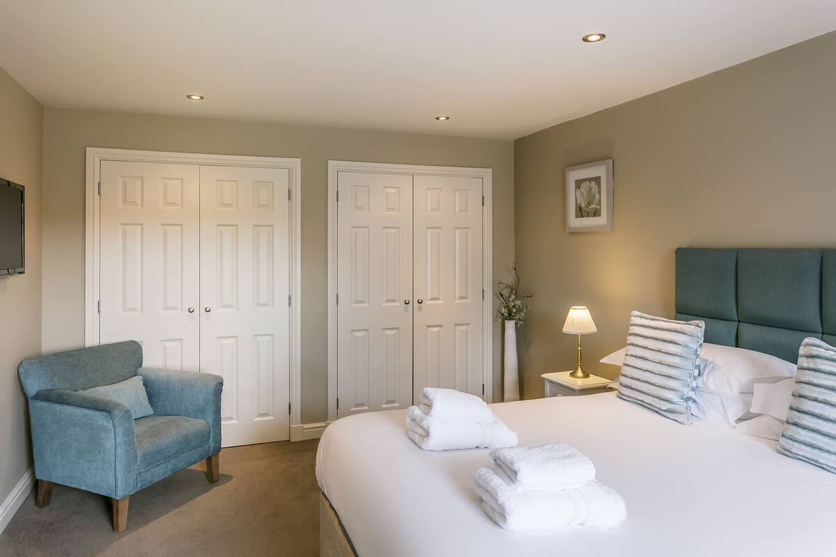A hotel room with a bed, a chair, a nightstand with a lamp, and two white doors.