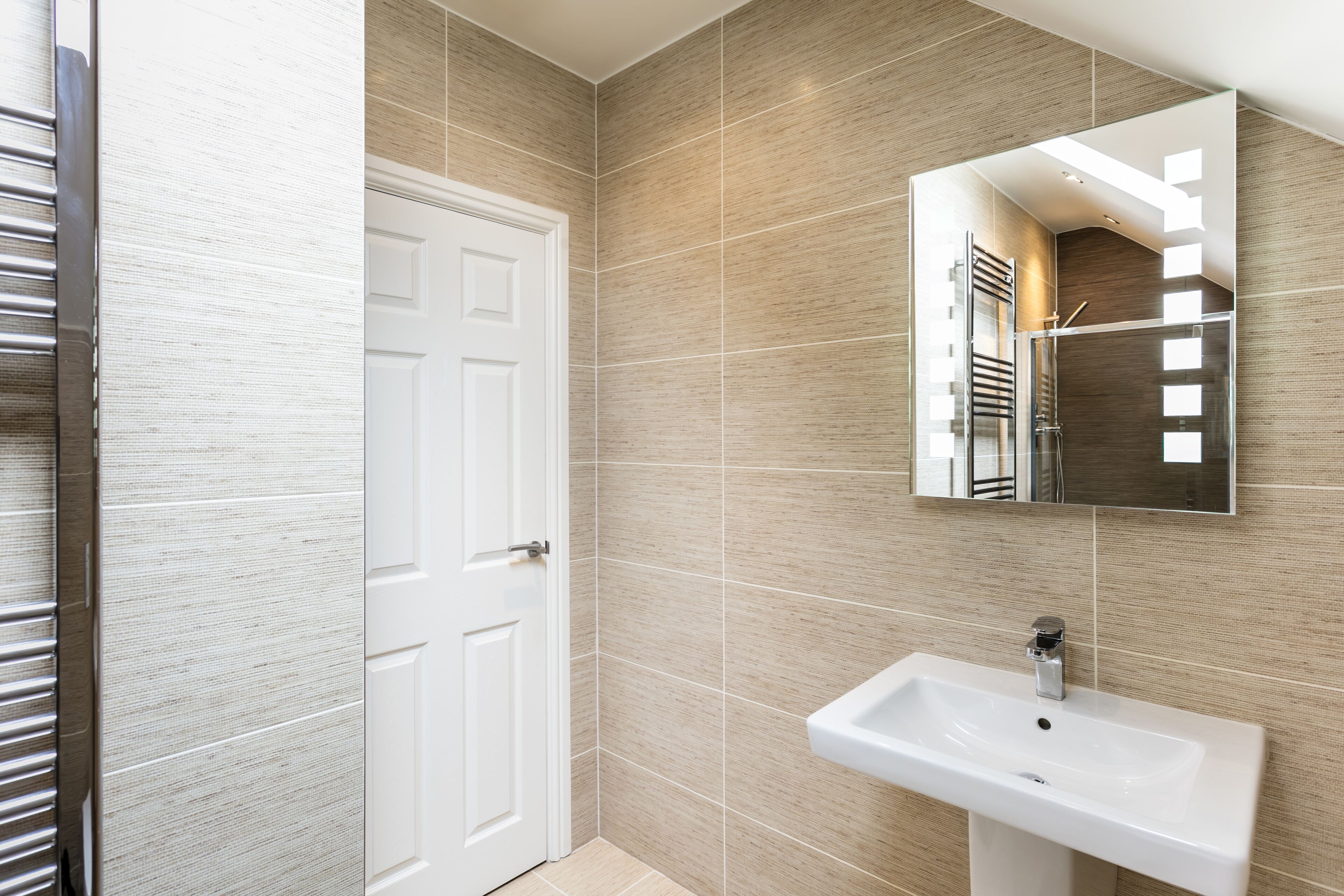 A modern bathroom with a white door, beige tiled walls, a large mirror, and a white sink.