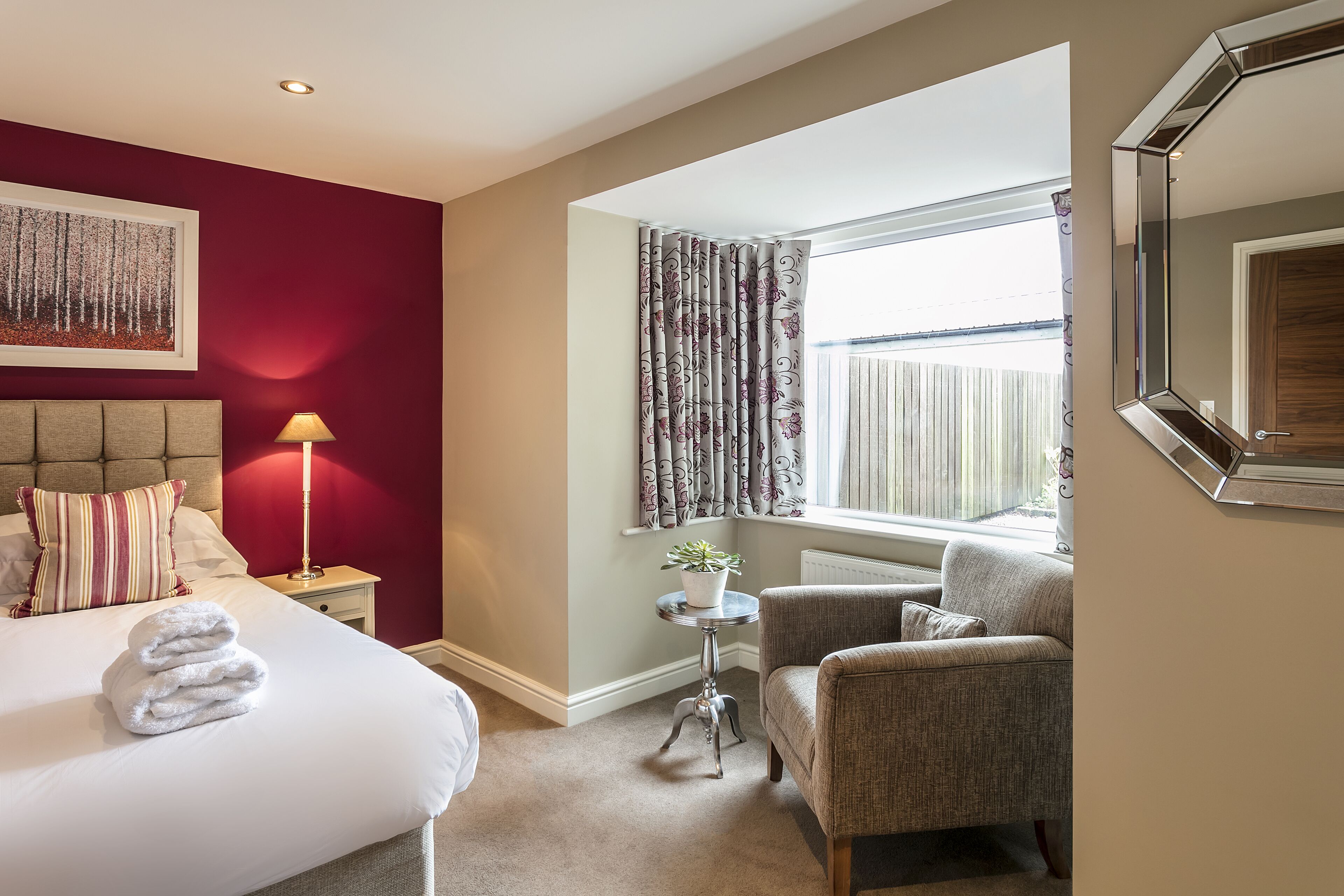 A hotel room with a bed, a chair, a small table with a lamp, a window with curtains, and a framed mirror.