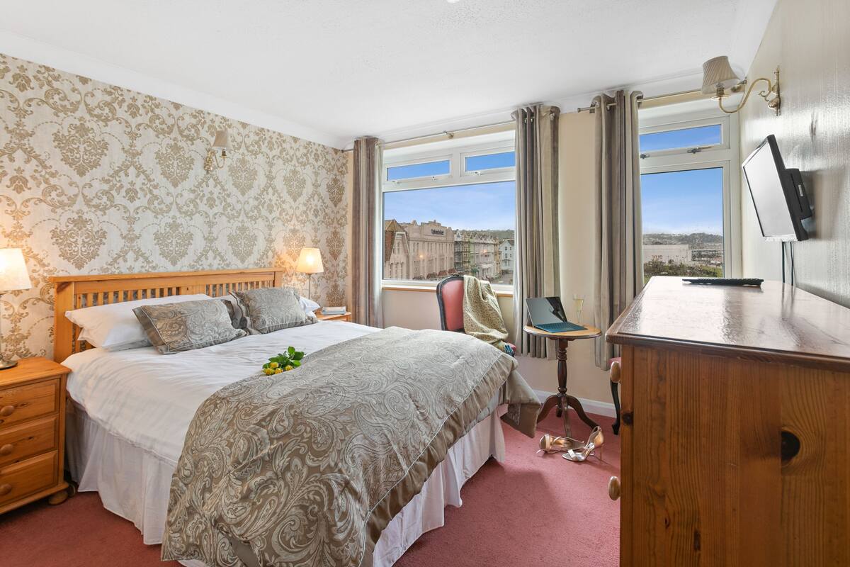 A hotel room with a large bed, a wooden dresser, a desk with a laptop, and a view of buildings through the window.