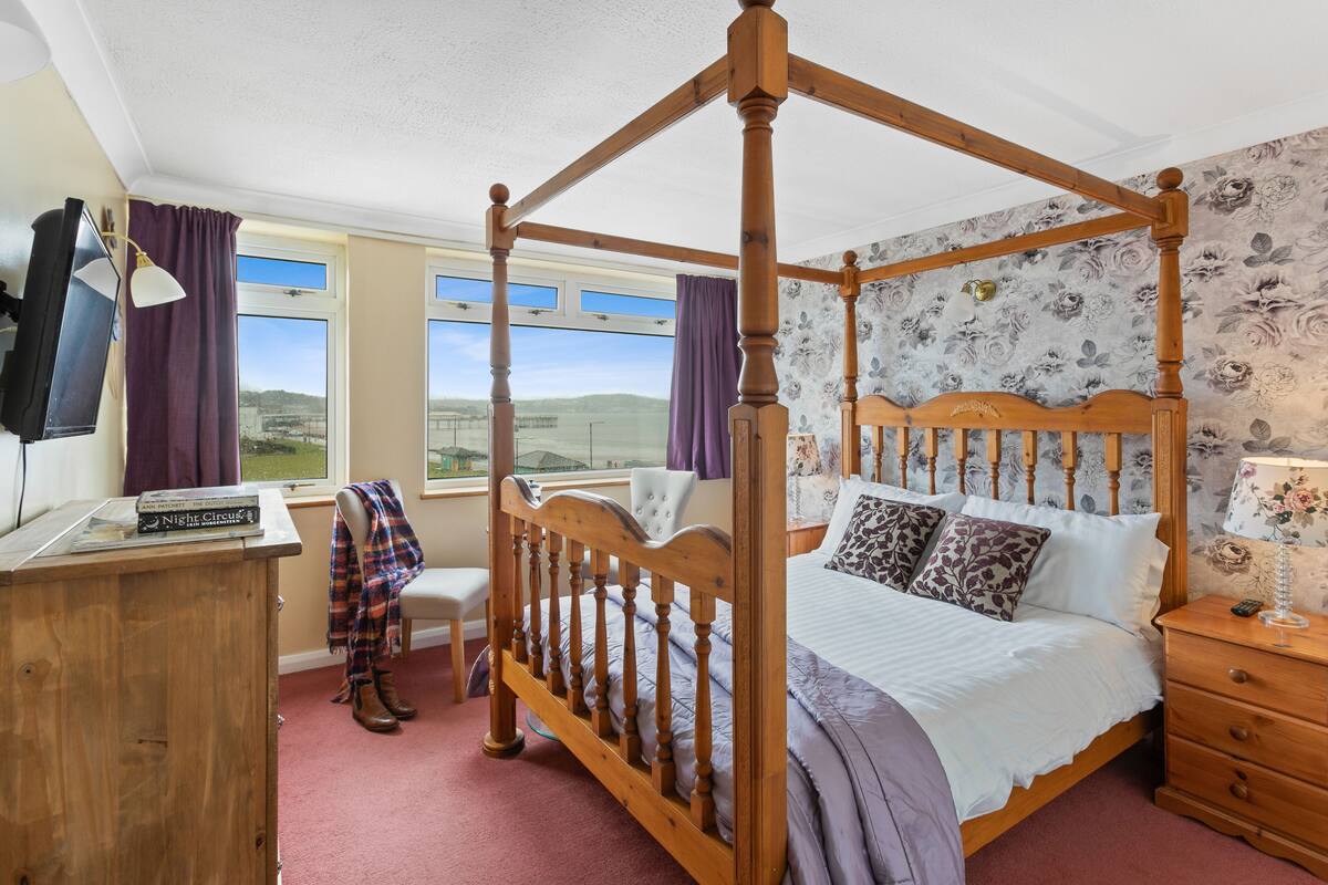 A four-poster bed with a headboard, a bedside table, a nightstand, a desk with a lamp, a chair, and a view of the outdoors through large windows.